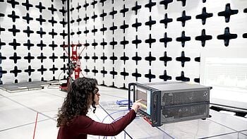 Engineer in a Keysight EMC anechoic test chamber, pointing at a rack instrument displaying a live RF spectrum trace, with pyramidal RF-absorber panels lining the chamber walls.